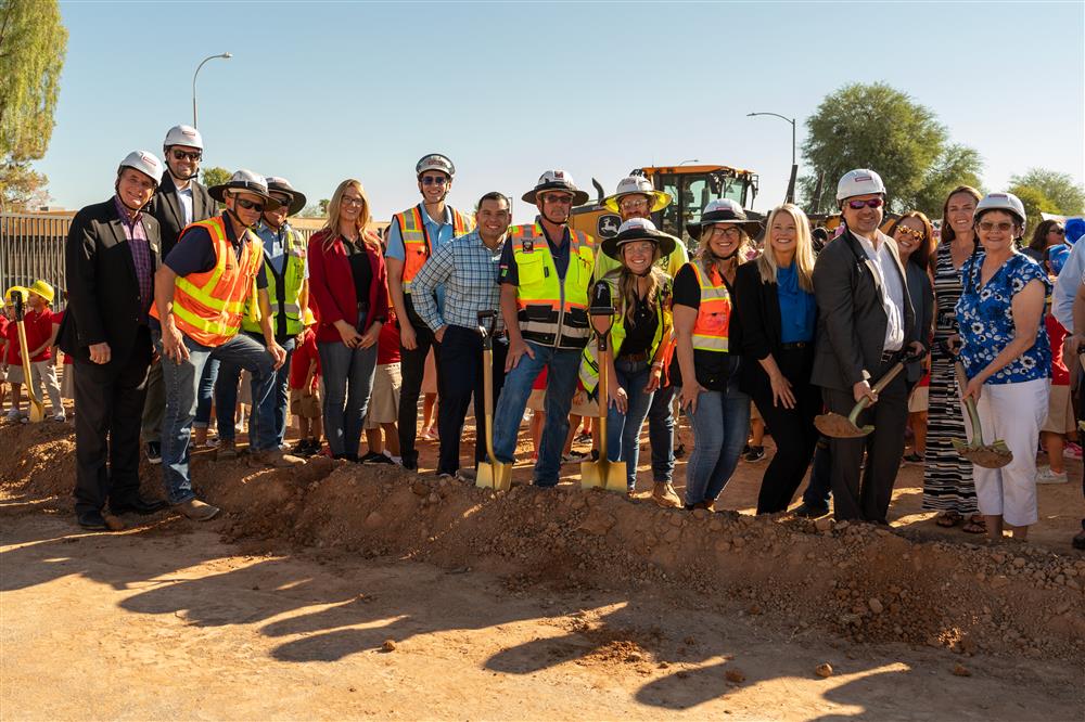 People dressed in hard hats and safety vests use shovels to dig into the ground